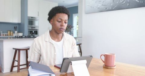 Young man working remotely on tablet at bright kitchen table with notebook and pink mug