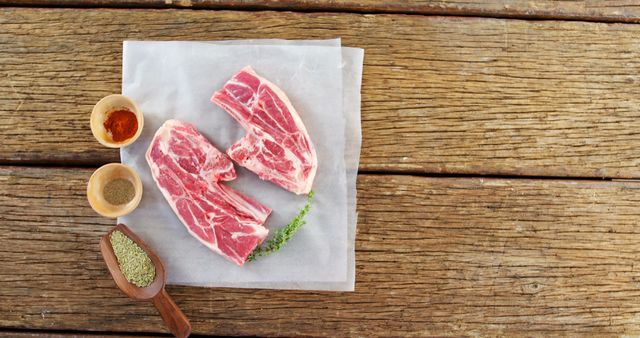 Raw lamb chops presented on parchment paper placed on a rustic wooden surface. Thyme, paprika, pepper and other seasonings are arranged next to the meat, ready for cooking. Ideal for use in culinary websites, recipe blogs, farm-to-table cooking promotions, or gourmet food advertising.