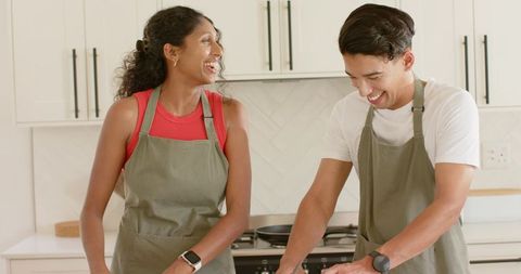 Joyful young couple cooking together in bright modern kitchen wearing matching aprons