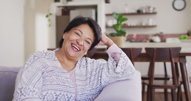 Senior woman sitting comfortably on a couch in a cozy living room, exuding happiness. Ideal for illustrating healthy aging, elderly lifestyle, retirement, and home comfort concepts. Perfect for use in health and wellness publications, retirement living advertisements, and family-oriented content.