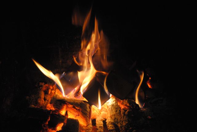 Close-up view of a fireplace with wood logs burning fiercely, generating flames and warmth in a dark environment. Ideal for illustrating concepts of home coziness, winter heating, fireplaces, and rustic warmth. Can be used in materials related to home improvement, interior design, and fire safety.