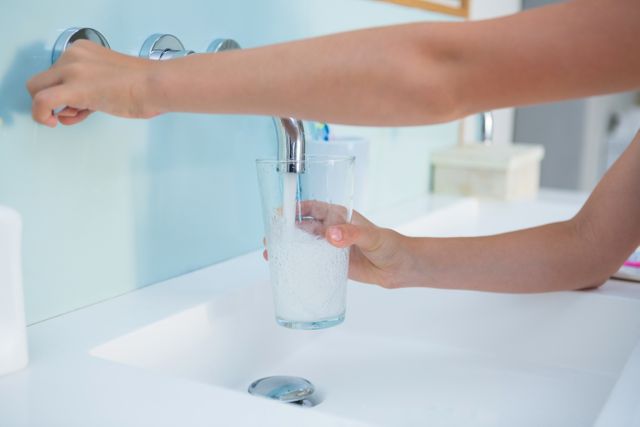 Hands filling a glass with water from a bathroom sink faucet. Ideal for illustrating concepts of hygiene, daily routines, hydration, and healthy living. Useful for articles, blogs, and advertisements related to home life, health, and wellness.