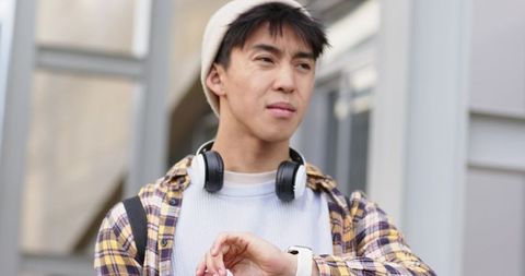 Young asian man checking smartwatch outside urban campus with headphones and backpack