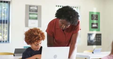 African American teacher guiding elementary student using laptop in bright modern classroom