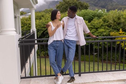 Diverse couple enjoying morning on balcony holding glass overlooking lush garden and hills