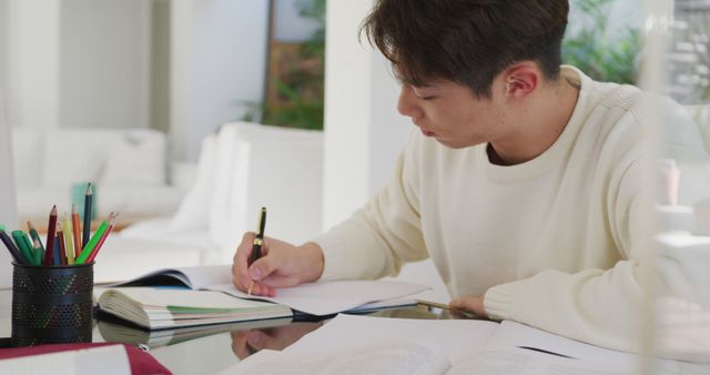 Young man is studying and writing notes at home. He sits at a desk filled with books and writing utensils, showcasing a typical academic and focused environment. This can be used for educational content, study tips, online courses, academic blogs, and motivational posts about education and productivity.