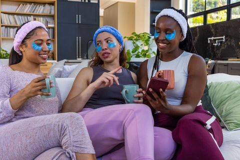 African american women enjoying cozy skincare day on sofa sharing coffee and smartphone