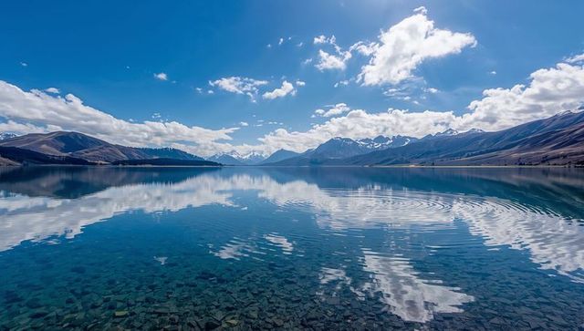 Crystal-clear alpine lake reflecting snow-capped peaks, blue sky, rocky shallow water