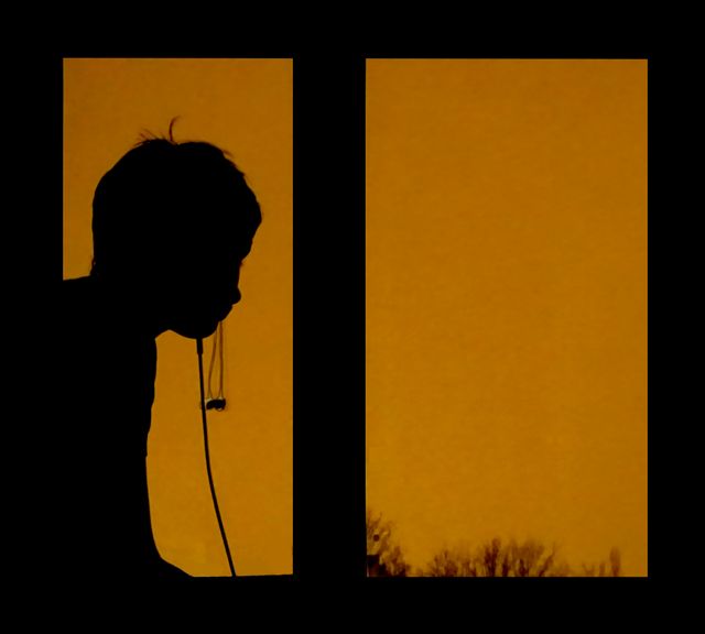 Silhouette of person leaning by a window during sunset with headphones around their neck. The orange sky contrasts against the dark figure, creating a tranquil and contemplative scene. Suitable for themes of relaxation, solitude, reflection, music, and evening time.