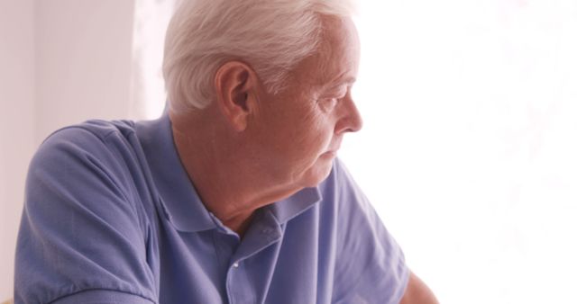 Elderly man deep in thought while bathed in natural light, wearing a blue polo shirt. Perfect for themes related to aging, retirement, contemplation, and celebrating the wisdom of elders. Great for use in lifestyle articles, health and wellness content, and advertisements targeting senior living or retirement communities.