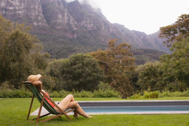 Woman in swimwear lounging on a sun lounger near a pool with a scenic mountain view. Ideal for use in travel brochures, vacation advertisements, wellness and relaxation promotions, and lifestyle blogs.