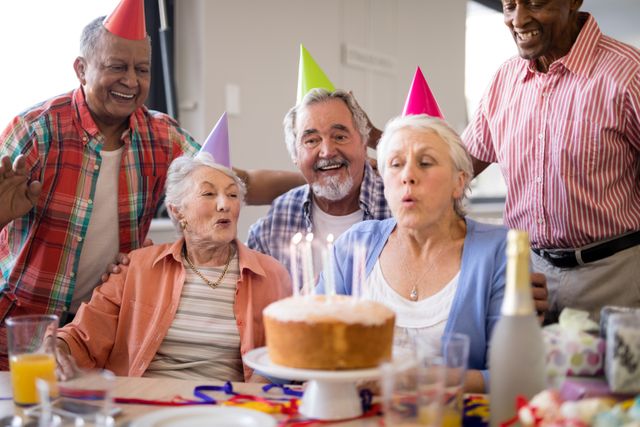 Senior woman blowing candles on birthday cake surrounded by friends in a nursing home. All are wearing party hats and smiling, creating a joyful and festive atmosphere. Perfect for use in advertisements for senior living communities, birthday celebration themes, and promoting elderly care services.