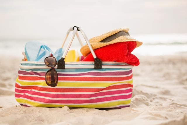 Colorful striped beach bag filled with summer essentials like a towel, hat, and sunglasses, placed on sandy beach. Ideal for promoting summer vacations, beach holidays, travel destinations, and leisure activities.