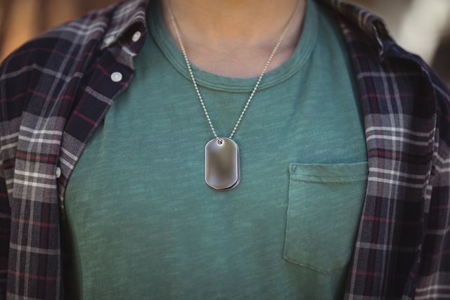 Midsection of a man wearing a dog tag pendant over a green shirt and plaid shirt. Ideal for use in fashion blogs, jewelry advertisements, casual wear promotions, and lifestyle articles.