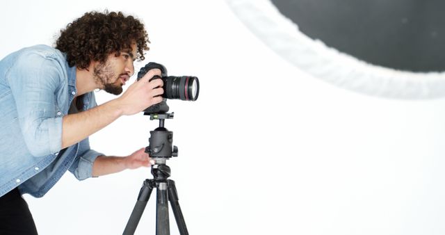 Professional photographer with curly hair adjusting camera on tripod in a studio setting. This can be used for content related to photography, professional work, studio sessions, photographic equipment, or photography tutorials. Ideal for articles, promotional material for photography courses, or marketing content for camera and tripod manufacturers.