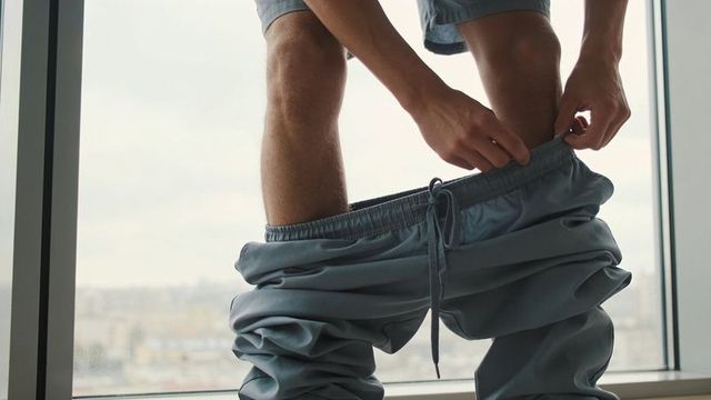 Man pulling down gray sweatpants beside large city window, candid intimate moment