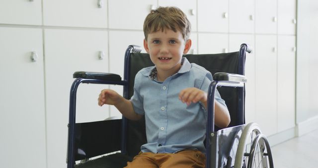 Image of a young boy smiling while sitting in a wheelchair in an indoor setting. The boy is wearing a light blue shirt and brown pants. There are cabinets in the background, suggesting a healthcare facility or hospital environment. This image can be used for themes related to disability, children's healthcare, positivity, rehabilitation, and inclusive lifestyle.