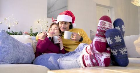 Couple cuddling on sofa holding mugs wearing santa hat and cozy holiday socks