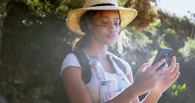 Young African American woman checks her phone outdoor. She's on a sunny hike, navigating with technology amidst nature.