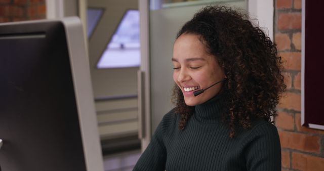 Biracial young professional wearing headset, working at computer. She has curly brown hair, light brown skin, and is wearing dark green turtleneck