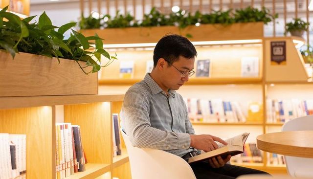 Asian man reading paperback in cozy bookstore lounge wearing glasses and checked shirt