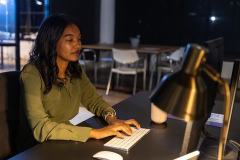 Working late in modern office professional woman typing on wireless keyboard by desk lamp