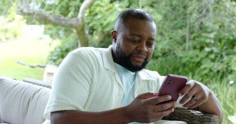 African american man relaxing on patio sofa using smartphone for messaging and leisure