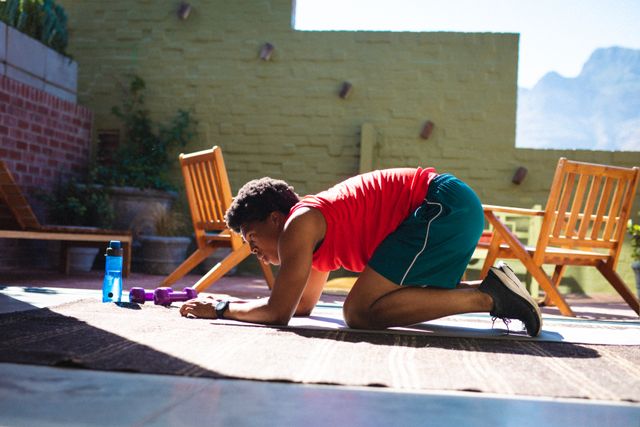 Side view of african american young man kneeling on exercise mat while exercising at home. unaltered, fitness, routine, stretching, sunlight and active lifestyle concept.
