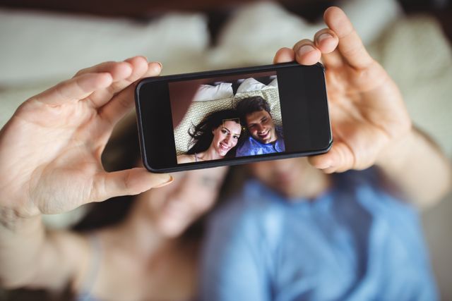Romantic couple lying on bed, taking a selfie with a smartphone. Ideal for use in articles or advertisements about relationships, love, modern technology, and lifestyle. Perfect for social media campaigns, blogs, and websites focusing on romance, intimacy, and young adult life.