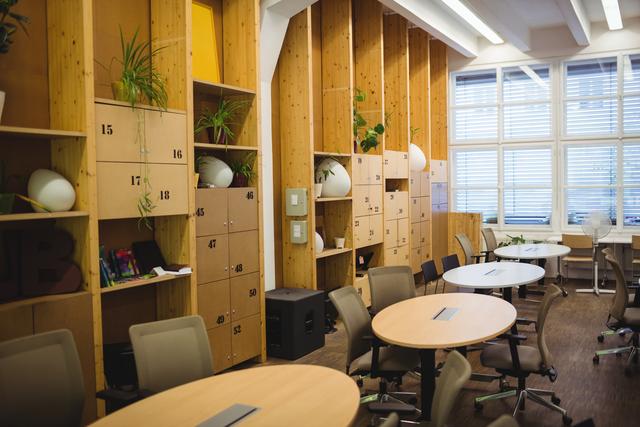 Modern empty office workspace featuring wooden lockers, round desks, and ergonomic chairs. Ideal for illustrating contemporary office design, professional work environments, or coworking spaces. Suitable for business presentations, office furniture advertisements, or articles on modern workplace trends.