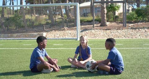 Diverse children sitting on soccer field near goal wearing matching blue kits holding ball