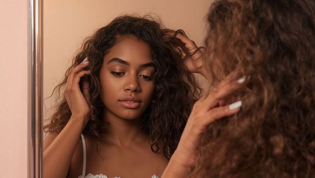 Young woman adjusting curly hair in chrome mirror, grooming routine, white lace camisole