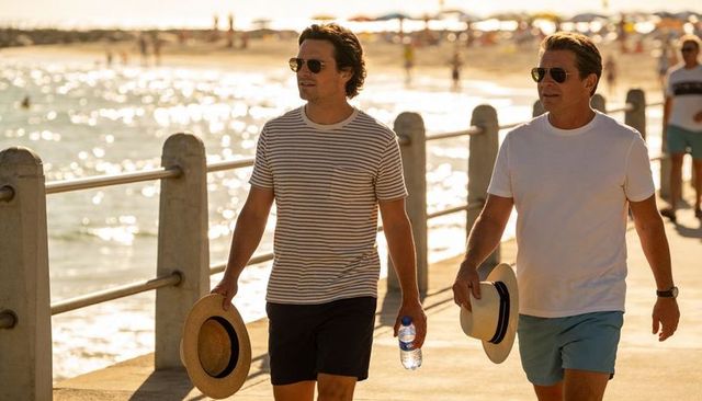 Two men walking on sunlit seaside promenade holding straw hats and water bottle