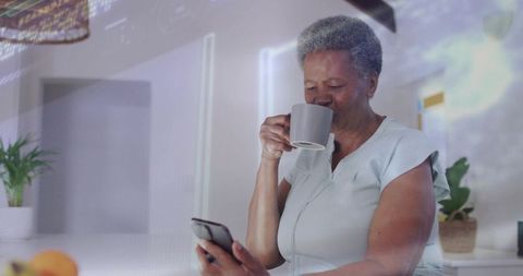 Senior woman enjoying morning coffee while checking smartphone in modern kitchen