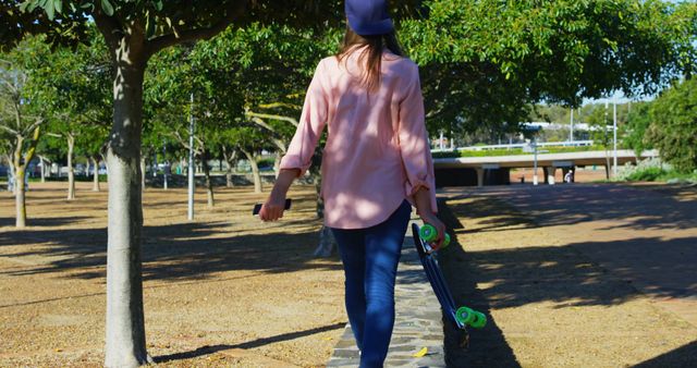 A young woman walks in a park on a sunny day with a skateboard in hand, dressed in casual clothing and a hat. This image is perfect for promoting outdoor activities, casual lifestyle, youth-focused content, or clothing brands.