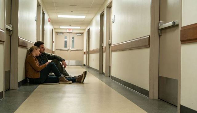 Young couple sitting, leaning on wall in clinic corridor using smartphone offering comfort