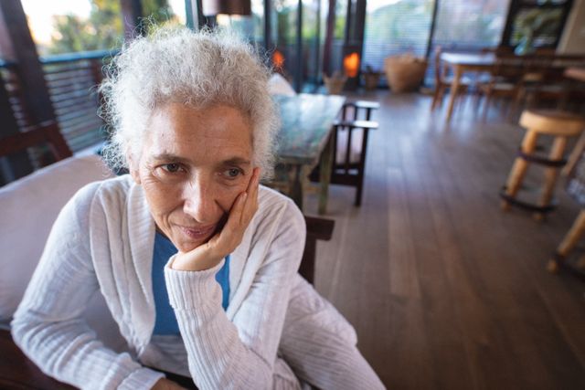 This image depicts a thoughtful senior woman sitting alone in a cozy home environment. It can be used in articles or advertisements related to retirement, aging, mental health, and senior living. The natural light and serene setting make it suitable for promoting peaceful and relaxed lifestyles for the elderly.