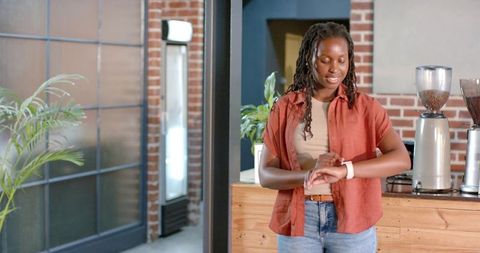 African woman checking smartwatch behind cafe counter with espresso grinders and brick wall