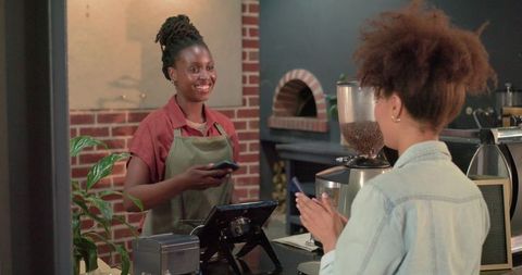 Smiling female barista exchanging mobile payment with customer at cozy brick cafe counter