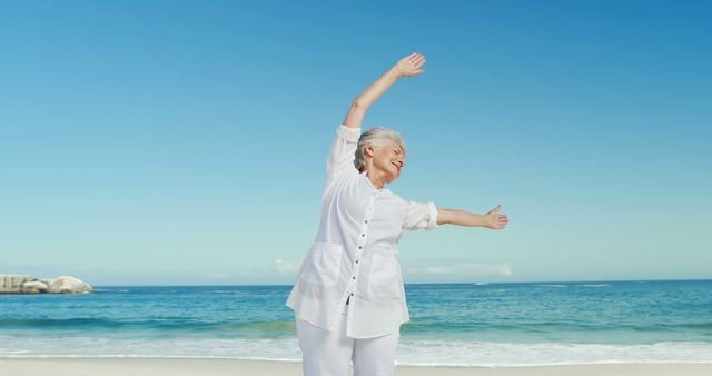 Senior woman enjoying stretching exercises on a beach with a calm ocean and clear blue sky in the background. Perfect for use in lifestyle, wellness, travel, and senior living advertisements, promoting healthy and active aging experiences.