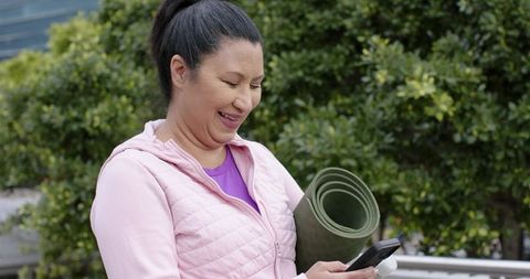 Mature asian woman checking smartphone while holding rolled yoga mat in urban greenspace