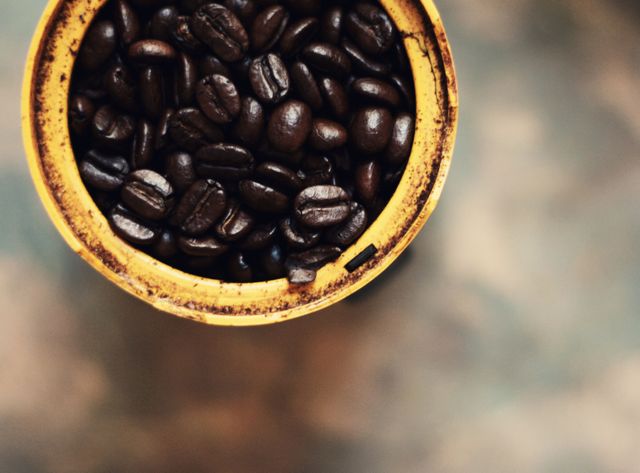 Close-up view of dark, roasted coffee beans inside a rustic, yellow container on an earthy background. Ideal for illustrating concepts related to coffee preparation, gourmet drinks, or rustic kitchen elements in blogs, websites, magazines, and advertisements.