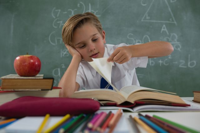 Young boy in school uniform reading a book at his desk in a classroom. Chalkboard with mathematical equations in the background. Apple and school supplies on the desk. Ideal for educational materials, school advertisements, and learning resources.