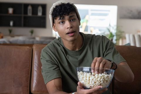 African american teen watching tv on sofa eating popcorn in cozy modern living room