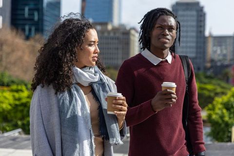 Diverse young couple walking, chatting and holding takeaway coffee against city skyline