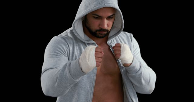 A young, athletic Middle Eastern man in a hoodie stands ready with his fists wrapped, suggesting he is a boxer or martial artist, with copy space. His intense gaze and poised stance convey determination and focus.