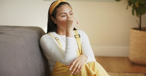 Asian woman sitting contemplative on floor leaning on sofa in cozy minimalist living room