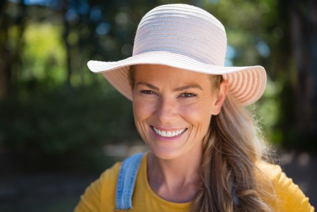 Woman smiling while wearing a sun hat outdoors on a sunny day. Ideal for use in advertisements, lifestyle blogs, summer promotions, and articles about outdoor activities, happiness, and fashion.