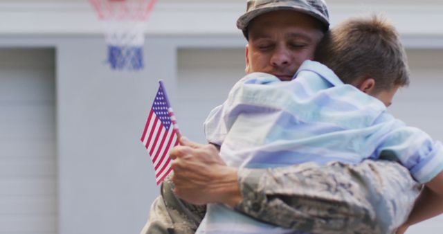 Happy caucasian male soldier hugging his smiling son holding flag in garden outside their house. soldier returning home to family.