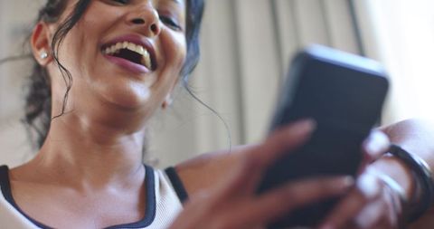 Cheerful woman laughing while using smartphone at home with casual tank top and jewelry
